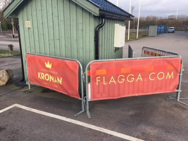 Röd reklambanderoll med Flaggfabriken kronan och texten flagga.com i gult på staket